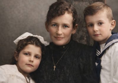 Das Foto zeigt eine Frau mit einem Mädchen im Vorschulalter und einem Jungen im Grundschulalter, ca. 1910er. Es ist nachcoloriert.